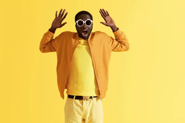 Scared man in sunglasses holding hands up on yellow background — Stock Photo, Image