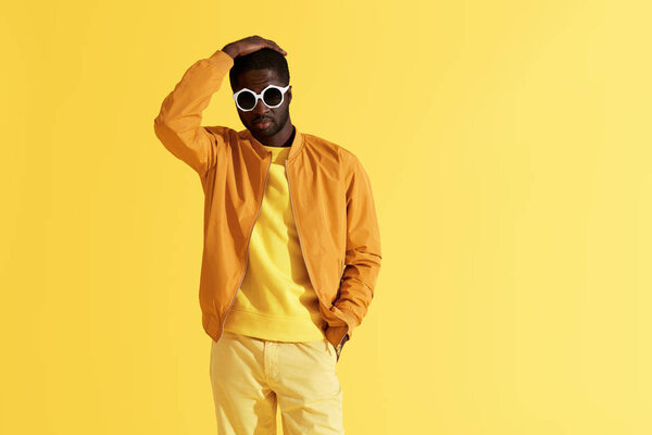 Fashion portrait of black man in stylish clothes and sunglasses on yellow background. Handsome african american male model in orange jacket posing in studio