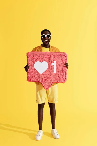 Like. Smiling black man with like icon, heart pinata on yellow — Stock Photo, Image