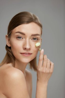 Evde yüz masajı için yeşim yüz silindiri kullanan kadın.