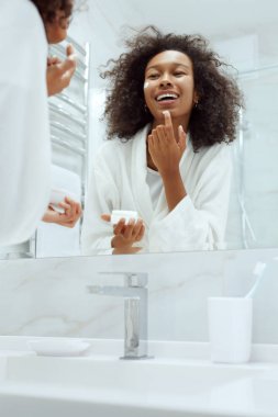 Cilt bakımı. Banyo aynasına bakan kadın yüz kremi sürüyor.