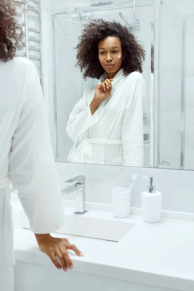 Skóra. Kobieta stosująca krem do twarzy patrząc w lustrze łazienki — Zdjęcie stockowe