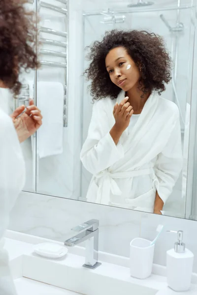 Skóra. Kobieta stosująca krem do twarzy patrząc w lustrze łazienki — Zdjęcie stockowe