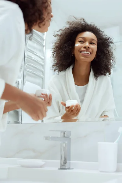 Cura della pelle. Donna che applica la crema viso guardando nello specchio del bagno — Foto Stock