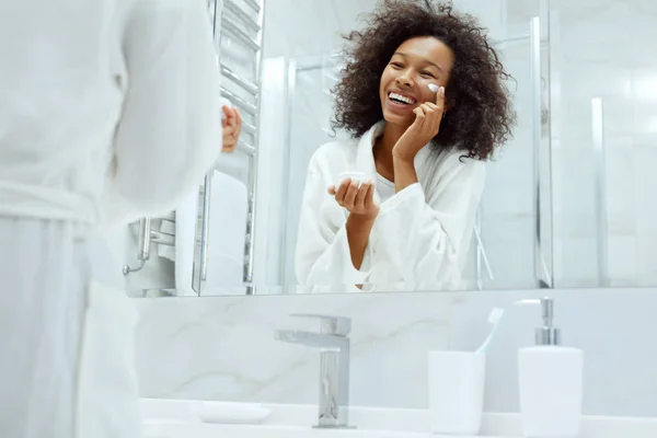 Cuidado com a pele. Mulher aplicando creme facial olhando no espelho do banheiro — Fotografia de Stock