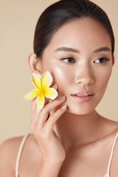 Beauty Face. Flower And Model Close Up Portrait. Beautiful Asian Woman With Plumeria Looking Away Against Beige Background. Girl With Natural Nude Makeup And Clean Healthy Skin After SPA Treatment.