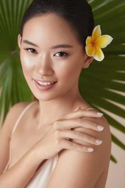 髪に花とパームの近くのモデル 熱帯植物の背景にあるアジアの女性の肩の肖像画に触れる公開 天然の有機化粧品を使用した後 美少女は水分補給肌を楽しむ — ストック写真