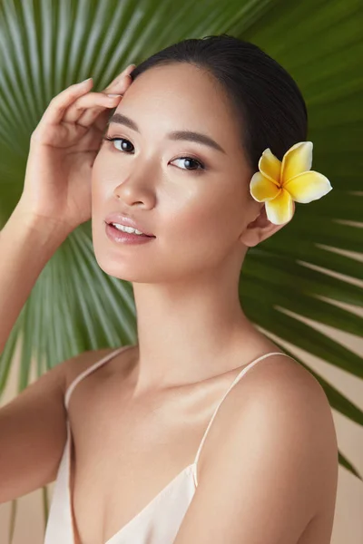 Woman Face. Leaf Of Palm Behind Asian Model. Beautiful Girl With Plumeria Flower In Hair Against Tropical Greenery Background. Organic Cosmetic And SPA Treatment For Healthy Hydrated Facial Skin.