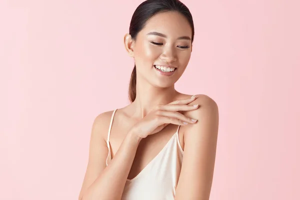 Beauty. Asian Woman Portrait. Ethnic Model With Smooth Skin Touches Her Shoulder And Smiling Against Pink Background. Confident Female Enjoying Her Perfect Body After Cosmetology Treatment.