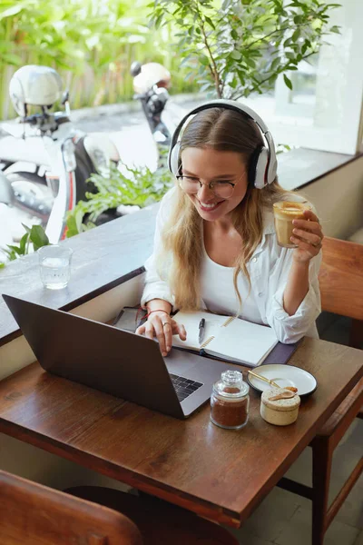 Girl At Coffee Shop. Woman Drinking Latte With Laptop And Headphones At Cafe. Beautiful Model In Glasses Working Or Studying Remotely At Comfortable Workplace On Vacation.