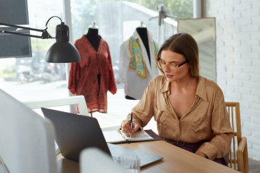 İş. Moda tasarımcısı stüdyoda çalışıyor. Çizim defterine bir şeyler yazan mutlu kadın. Modern yaratıcılık hobisi başlangıçtan başarıya. Kendi Atölyesinde Dizüstü bilgisayarı olan Serbest Çalışan Kadın.