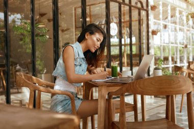 Çalış. Kahve dükkanında dizüstü bilgisayarı olan bir kadın. Cafe 'de kotlu Asyalı kız kitap yazıyor. Modern Dijital Teknolojilerle Uzaktan Çalışma veya Çevrimiçi Çalışmalar.