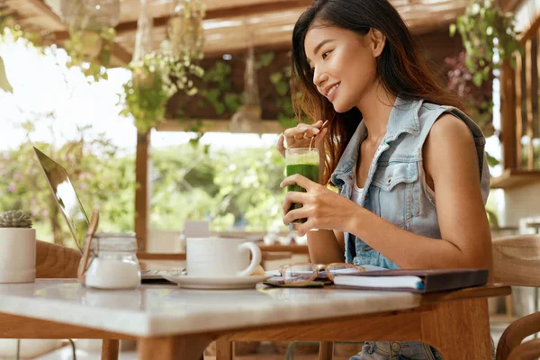 仕事だ 女はカフェでスムージーを飲む ラップトップにピッタリのジーンズ姿の幸せなアジアの女の子フレッシュなデトックスカクテルをお楽しみください デジタルノマドライフスタイルとオンライン作業のための現代技術 — ストック写真