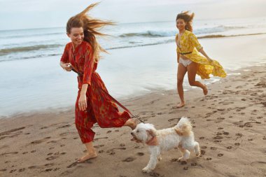 Kumsal. Kumsalda Köpeği Çıplak Ayakla Yürüyen Kızlar. Tropikal Okyanus 'ta yaz tatilinde evcil hayvanla bohem elbiseli moda kadınları. Şık görünüm ve dinlenme tesisinde rahat yürüyüş için Boho tarzı.