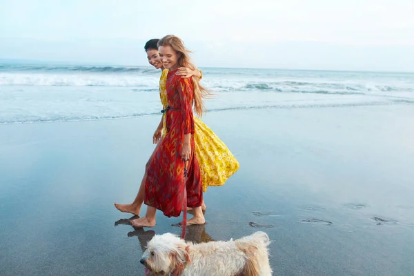 Dog-Friendly Beach. Women Walking With Pet On Sandy Coast And Enjoying Summer Vacation Near Ocean. Stylish Girls In Bohemian Dress Having Fun. Boho Clothing Style For Fashion Look On Resort.