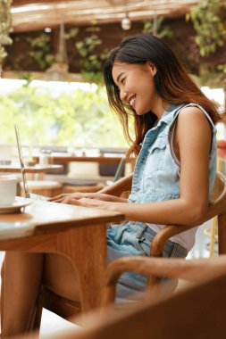Cafe 'de çalışıyorum. Online toplantı için dizüstü bilgisayar kullanan bir kadın. Kahve dükkanındaki kot elbiseli mutlu Asyalı kız. Konforlu İletişim için Modern Teknoloji ile Dijital Göçebe Yaşam Tarzı.