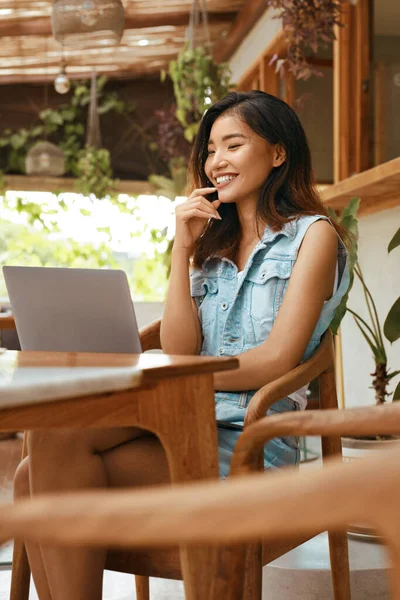 女性はカフェでオンライン会議を持っています テラスでラップトップを使用してカジュアル服で幸せなアジアの女の子と誰かとチャット 快適な職場でのリモートジョブや教育のための近代的な技術 — ストック写真