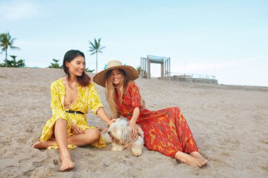 Kumsal. Kumsalda Köpekli Kadınlar. Hasır Şapkalı Bohem Giysili Moda Kızları Tropikal Sahilde Dinleniyor. Boho Modaya uygun görünüş ve yürüyüş stili.