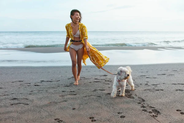 Dog-Friendly Beach. Woman Walking With Pet On Sandy Coast And Enjoying Summer Vacation Near Ocean. Fashion Girl In Bohemian Dress Having Fun. Boho Clothing Style For Fashionable Look.