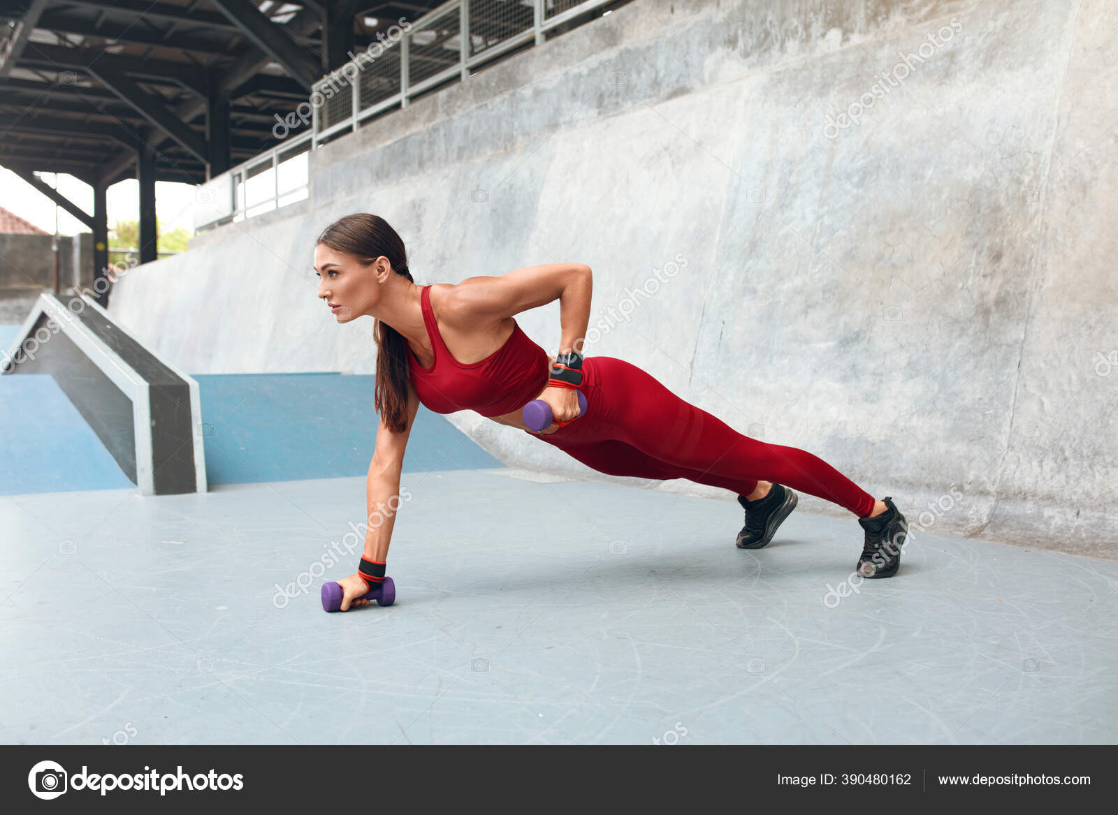Woman. Plank Workout With Dumbbells. Fitness Girl Training Outdoor For Strong Muscular Body. Female In Fashion Sportswear Lifting Hand With Sport Equipment. Intense Exercise Against Concrete Wall. Sto...