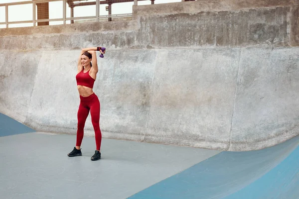 Edzés. Fitness lány emelés Dumbbells és csinál intenzív testmozgás. Sportos nő a divat sportruházat képzés ellen beton fal. Aktív városi életmód az erős izomzathoz. — Stock Fotó