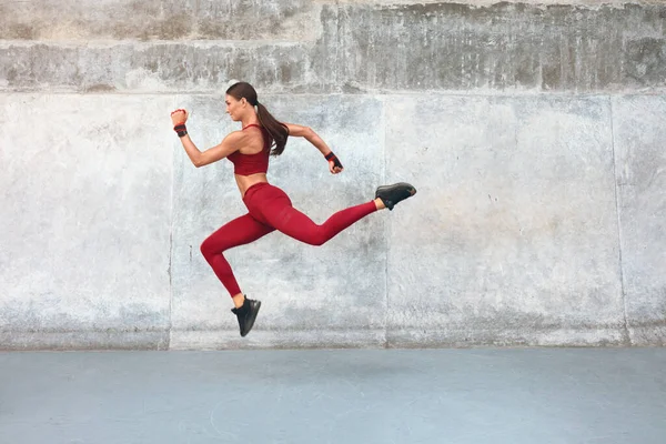 Fitness Girl Jumping. Venkovní cvičení proti betonové zdi na stadionu. Módní sportovní žena se silným sexy tělem v dynamické akční póze. Sport pro aktivní městské osoby. — Stock fotografie