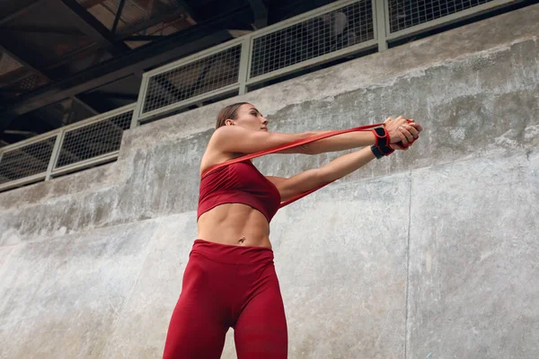 Ejercicios de mujer con banda de resistencia. Chica de fitness en el entrenamiento al aire libre en la ciudad. Sexy hembra en ropa deportiva de moda de entrenamiento contra la pared de piedra. Estilo de vida activo para un cuerpo muscular fuerte . —  Fotos de Stock