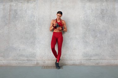 Spor salonu. Bir şişe Protein Shake ve direniş bandı olan fitness kızı duvarın yanında duruyor. Yoğun Kardiyo Çalışmaları Arasında Moda Giyen Sporcu Kadın. Şehir Yaşam Tarzı Olarak Açık Hava Egzersizi.