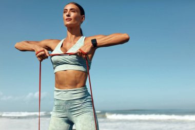 Plajdaki Fitness Kızı Portresi. Tropikal Okyanus Arkaplanı 'nda Direnç Döngüsü ile Esneme Çalışması. Güçlü kaslı vücutlu sporcu kadın yaz tatilinde moda spor kıyafetleri eğitimi alıyor..