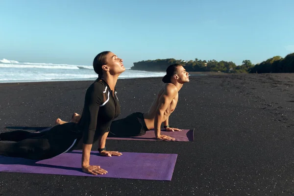 Deporte en la playa. Estiramiento de la pareja en el entrenamiento de ropa deportiva de moda. Hombre guapo y mujer sexy entrenando en Yoga Mat en la mañana. Estilo de vida activo en las vacaciones de verano en el mar tropical . — Foto de Stock