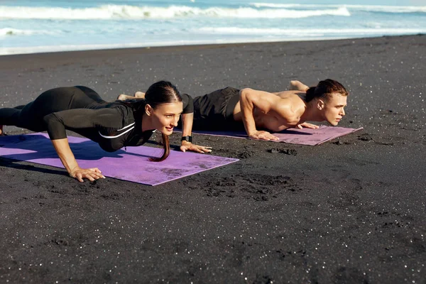 Un par de ejercicios en la playa. Hombre guapo y mujer sexy en ropa deportiva de moda haciendo flexiones en la esterilla de yoga por la mañana. Estilo de vida activo y deporte de recreación en las vacaciones de verano . — Foto de Stock