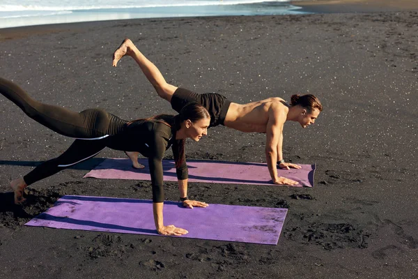 Strand. Frau und Mann trainieren morgens. Sportpaar in modischer Sportbekleidung macht Dehnübungen mit anheben des Beines auf der Yogamatte. Ausbildung im Sommerurlaub als Teil eines aktiven Lebensstils. — Stockfoto