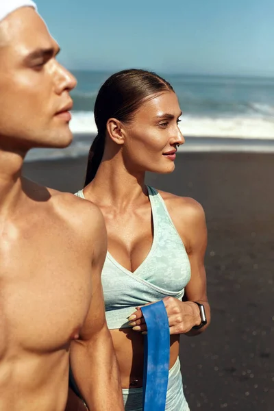 Retrato de pareja deportiva. Hombre guapo y mujer sexy posando contra el fondo del océano tropical. Entrenamiento de pareja con banda de resistencia en la playa. Vacaciones de verano activas como estilo de vida . —  Fotos de Stock