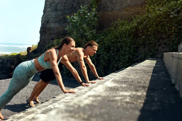 Deporte. Ejercicio de hombre y mujer en las escaleras. Un par de jóvenes deportistas en ropa de fitness de moda haciendo push-ups. Entrenamiento al aire libre para el cuerpo muscular fuerte. Estilo de vida activo en las vacaciones de verano . —  Fotos de Stock