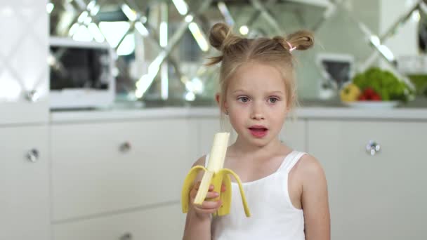 Дитина їсть їжу. Маленька дівчинка їсть банан на кухні — стокове відео