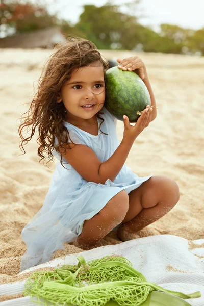 Дитина на пляжі Портрет. Мила маленька дівчинка з кучерявим волоссям тримає кавун. Щаслива дитина збирається поїсти на піщаному узбережжі. Органічні фрукти і натуральні вітаміни для здорового харчування на пікніку . — стокове фото