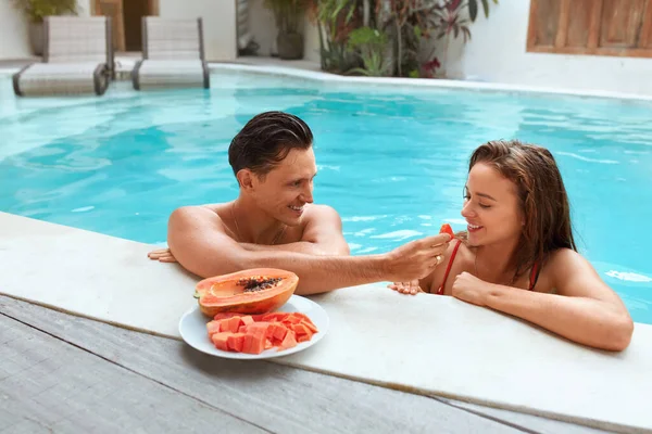 Par i kärlek vid poolen äta exotisk frukt. Stilig man och vacker kvinna njuter Papaya på sommarsemester. Man och kvinna på smekmånad eller romantisk helg på Tropical Villa. — Stockfoto