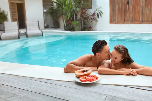 Ett par i poolen. Stilig man kysser vacker kvinna på turkost vatten bakgrund. Njuter av tropisk frukt på sommarsemester i Villa. Romantisk helg på Resort för positiva känslor. — Stockfoto