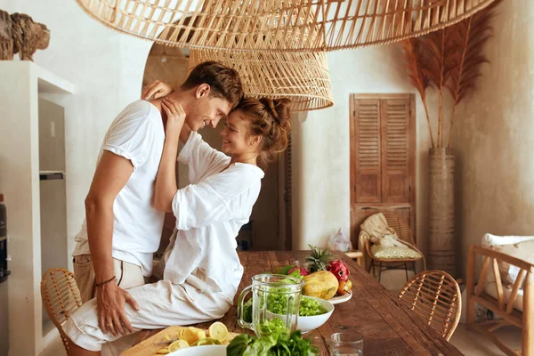 Couple romantique à la cuisine. Belle femme assise sur une table en bois et étreignant un homme debout. Amour et soins pour une relation heureuse comme mode de vie. Alimentation saine pendant les vacances d'été à la villa tropicale. — Photo