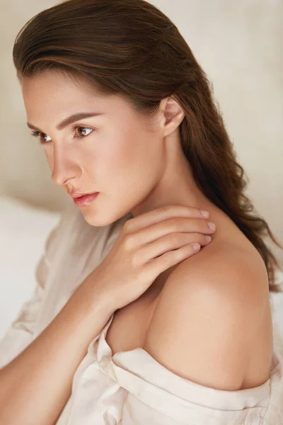 Belleza. Mujer con maquillaje natural y piel facial hidratada brillante. Retrato de hermosa modelo tocando hombro desnudo y mirando hacia otro lado. — Foto de Stock