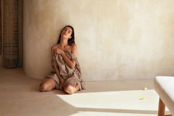 Kvinnan. Porträtt av vacker modell sitter nära väggen på terrassen. Förförisk brunett med perfekt slät solbränd hud som lägger sig på golvet med blommor i solljus och tittar på kameran. — Stockfoto