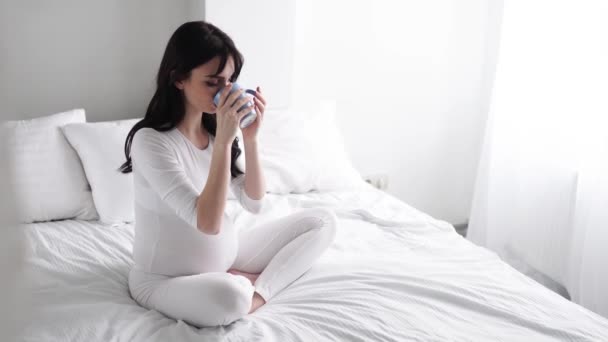 Gravid ung kvinna dricker te och sitter på sängen hemma — Stockvideo