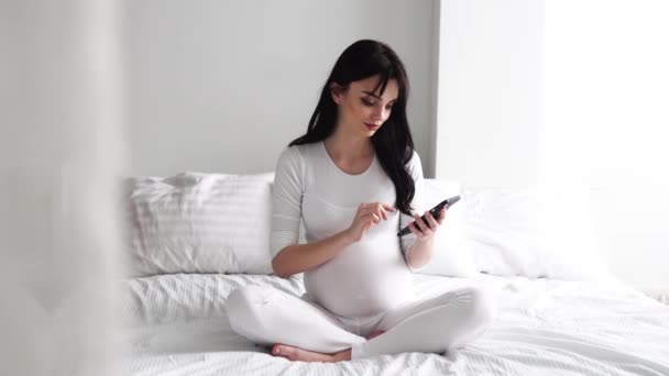 Femme enceinte avec téléphone portable assis sur le lit dans la chambre à coucher — Video