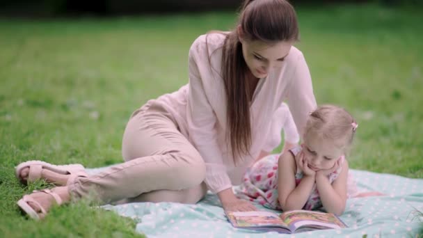 Jovem mãe e filha lendo livro deitado na grama no parque — Vídeo de Stock