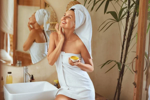 Mujer después del baño. Retrato de modelo feliz con toalla después de la ducha. Joven hembra que aplica crema antienvejecimiento o máscara en la piel facial. Producto de belleza natural para el tratamiento del SPA en el hogar. — Foto de Stock