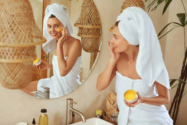 Vrouw die Clay Mask toepast. Verpakt in bad handdoek Model kijken naar spiegel en het gebruik van huidverzorging product voor Derma. Morning Routine met natuurlijke cosmetica voor een perfecte look. — Stockfoto