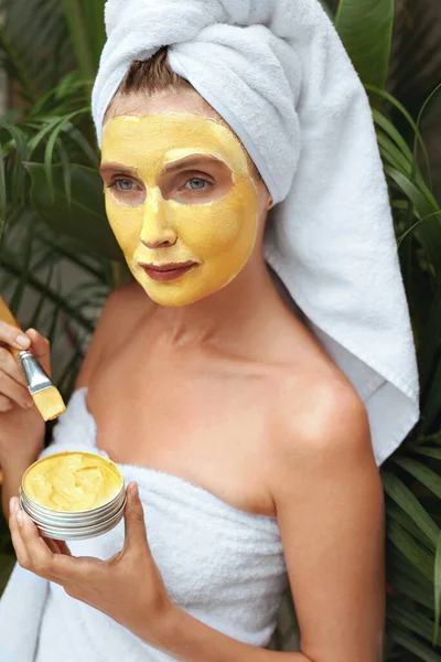 Huidverzorging. Een vrouw met een geel kleimasker op haar gezicht. Vrouw in badhanddoek met borstel voor het aanbrengen van Beauty Product met kurkuma. SPA behandeling en anti-veroudering therapie voor perfecte gezichtsbehandeling Derma. — Stockfoto