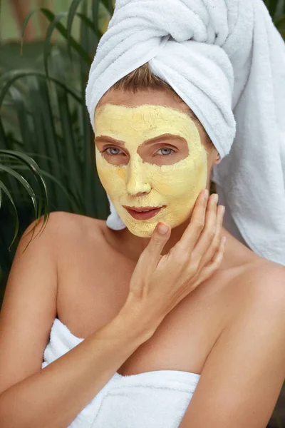 Vrouw in badhanddoek met kurkuma masker op het gezicht. Close-up Portret van jonge vrouw aanraken gezichtshuid bedekt met schoonheid product. Model na SPA behandeling tegen tropisch groen. — Stockfoto