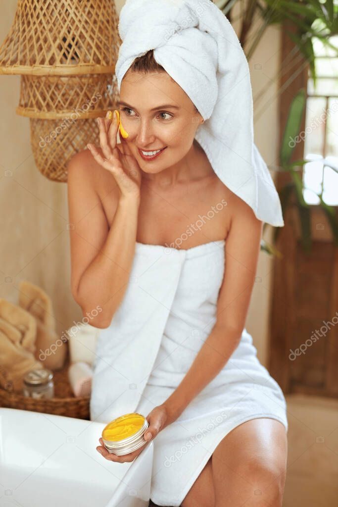 Skin Care. Woman Applying Yellow Clay Mask On Face. Model In Bath Towel Sitting In Bathroom And Using Beauty Product With Turmeric. SPA Treatment And Anti-Aging Therapy For Perfect Facial Derma.
