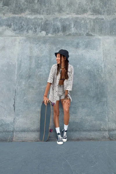 Dziewczyna na łyżwach w skateparku. Portret kobiety hipstera w luźnym stroju z deskorolką. Nastolatek patrzący na betonową ścianę w Skate Park. Styl subkultury miejskiej i sport letni. — Zdjęcie stockowe